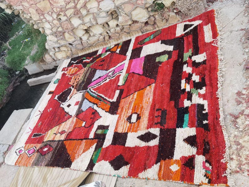 May include: A colorful Moroccan rug with geometric patterns in shades of red, orange, white, and black. The rug has a fringed edge and is made of wool.