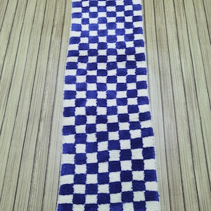 Puede incluir: Una alfombra larga y rectangular a cuadros azules y blancos con flecos en la parte inferior. La alfombra está hecha de un material suave y texturizado y se coloca sobre una superficie de madera. El patrón de cuadros está formado por cuadrados azules y blancos alternos.