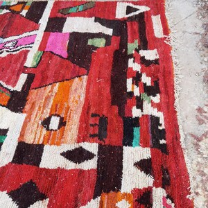 May include: A colorful Moroccan rug with geometric patterns in shades of red, orange, white, black, and green. The rug has a frayed edge and is made of wool.