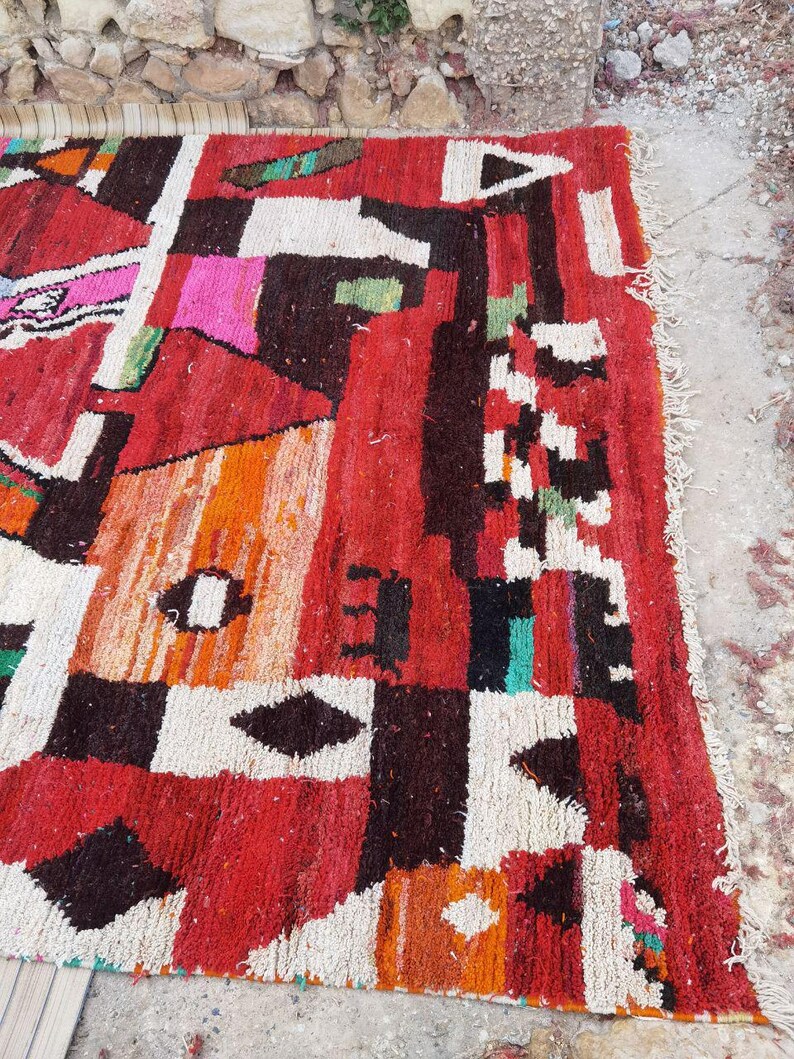 May include: A colorful Moroccan rug with geometric patterns in shades of red, orange, white, black, and green. The rug has a frayed edge and is made of wool.