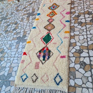 Puede incluir: Alfombra rectangular de color crema con un patrón geométrico. La alfombra presenta formas de diamante en varios colores, incluyendo verde, azul y rosa, conectadas por líneas en zigzag. La alfombra tiene flecos en el borde inferior.