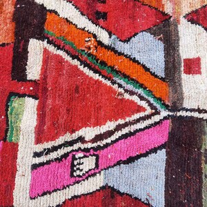 May include: A close-up of a colorful rug with geometric patterns in shades of red, orange, pink, blue, green, and white. The rug is made of woven wool and has a textured surface.