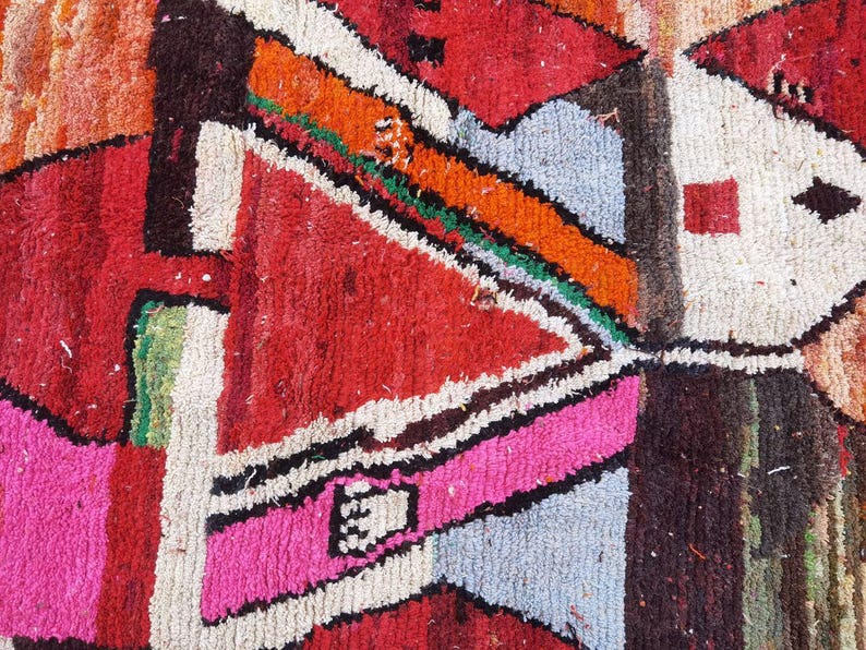 May include: A close-up of a colorful rug with geometric patterns in shades of red, orange, pink, blue, green, and white. The rug is made of woven wool and has a textured surface.
