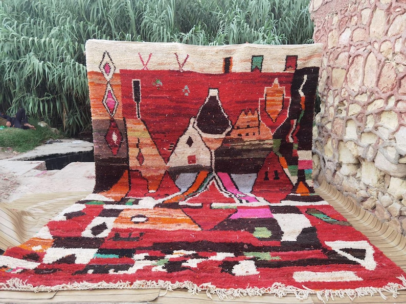 May include: A colorful Moroccan rug with geometric patterns in shades of red, orange, black, white, and green. The rug features a unique design with abstract shapes and a central motif of a stylized building.