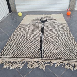May include: A black and white Moroccan rug with a geometric pattern and fringe. The rug has a central design that resembles a stylized arrow.