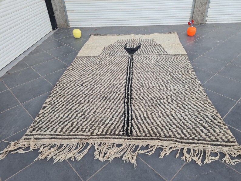 May include: A black and white Moroccan rug with a geometric pattern and fringe. The rug has a central design that resembles a stylized arrow.