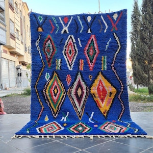 May include: A vibrant blue rug with a geometric pattern. The rug features diamond shapes in red, yellow, green, and white, with a fringe along the bottom edge. The rug is displayed outdoors.