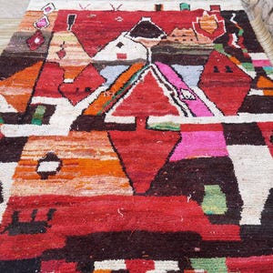 May include: A colorful Moroccan rug with geometric patterns in shades of red, orange, white, black, and green. The rug features a central diamond shape with smaller geometric shapes surrounding it.