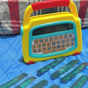 Puede incluir: Un juguete amarillo y azul con un asa roja y un teclado con el alfabeto. Varias etiquetas de madera verdes con texto están esparcidas alrededor del juguete. El texto en las etiquetas incluye "YOU'RE WELCOME" y "LOU'S THIGH BURN".