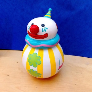 Può includere: Una stravagante statuetta a forma di clown con una testa bianca, un naso rosso e un colletto blu e rosa. Il corpo è a righe gialle e bianche con accenti floreali verdi. Il clown ha un piccolo cappello a righe.