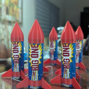 May include: Several toy rockets in red, white, and blue. Each rocket features a red cone, red fins, and a blue body with the text "THE BIG ONE" in a yellow burst design. The rockets are displayed on a brown surface.