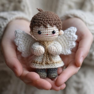 Puede incluir: Una muñeca de ángel de ganchillo hecha a mano con cabello castaño, ojos negros pequeños y una sonrisa roja. La muñeca tiene alas blancas y viste un atuendo color crema con ribete dorado. La muñeca se sostiene en manos abiertas.