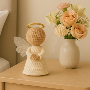 Puede incluir: Una figura de ángel de ganchillo hecha a mano con un halo dorado y alas delicadas. El ángel viste un vestido color crema y tiene la cabeza y las manos de color marrón claro. Un pequeño jarrón con rosas color melocotón y otras flores está cerca.