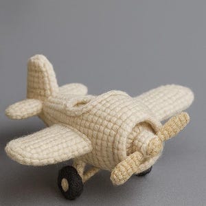 Op de afbeelding: Een gehaakt speelgoedvliegtuig in crèmekleur met een propeller en zwarte wielen. Het handgemaakte speelgoed heeft vleugels en een staart, tegen een grijze achtergrond.
