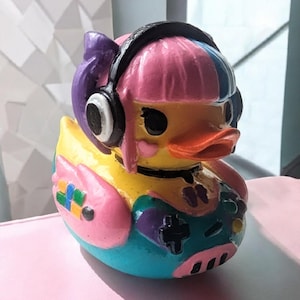 以下が含まれることがあります： ゲームをテーマにした手描きのカラフルなアヒルの置物。アヒルはピンク、紫、青の髪、黒のヘッドセット、胸にはコントローラーのデザインがあります。アヒルは黄色、ピンク、青、緑です。