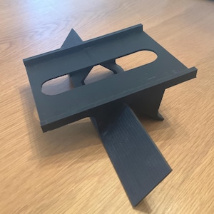 May include: A black plastic stand with two slots for holding a device, such as a phone or tablet. The stand is designed to prop up the device at an angle.