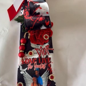 Può includere: Un nastro commemorativo con un fiocco rosso e bianco in alto, nastri a motivi bianchi e neri, una rosa di stoffa rossa, una spilla da balia con perline rosse e un ciondolo color argento Dior. Il nastro mostra foto e il testo LONG LIVE.