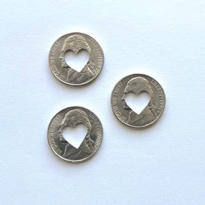 May include: Three silver nickels with heart-shaped cutouts. Each coin features the profile of a man and the words "LIBERTY" and "IN GOD WE TRUST". The years 1991, 1999, and 2000 are visible on the coins.