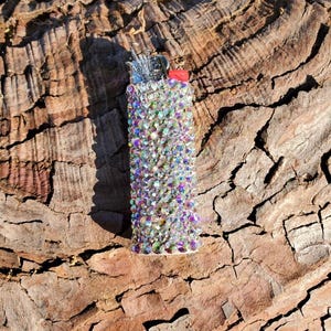 May include: A bedazzled lighter covered in iridescent rhinestones. The lighter has a red button and silver metal components. The rhinestones reflect a rainbow of colors, creating a sparkling effect. The lighter is set against a textured, brown background.