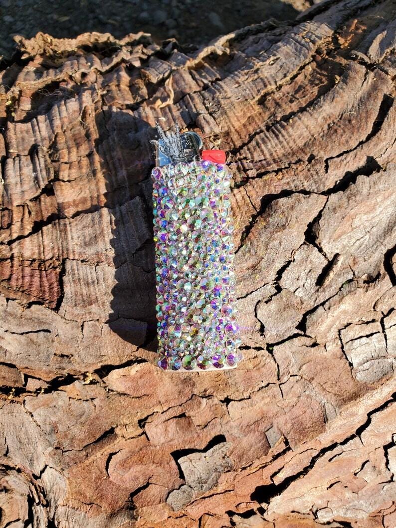 May include: A bedazzled lighter covered in iridescent rhinestones. The lighter has a red button and silver metal components. The rhinestones reflect a rainbow of colors, creating a sparkling effect. The lighter is set against a textured, brown background.