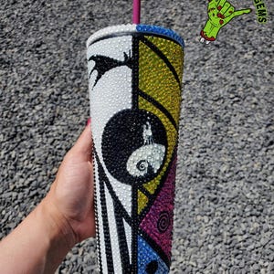 May include: A white, black, yellow, blue, and pink tumbler with a rhinestone design featuring Jack Skellington from The Nightmare Before Christmas. The tumbler has a black and white striped design on the left side and a geometric design on the right side. The tumbler has a clear lid and a pink straw.