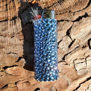 May include: A blue rhinestone-covered lighter with the text "Wicked Gems" on the side. The lighter is a standard size and shape, with a metallic top. The rhinestones are a deep blue color and cover the entire body of the lighter.