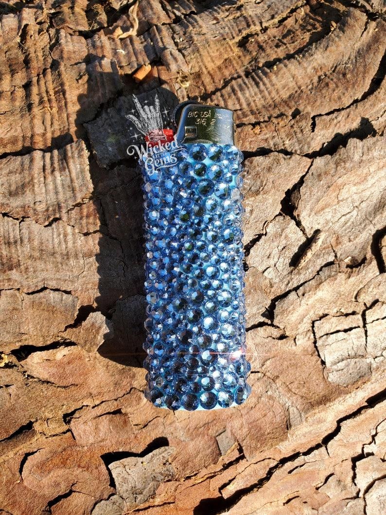 May include: A blue rhinestone-covered lighter with the text "Wicked Gems" on the side. The lighter is a standard size and shape, with a metallic top. The rhinestones are a deep blue color and cover the entire body of the lighter.