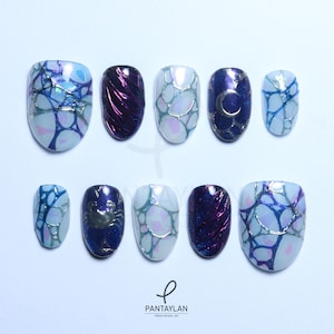 May include: A set of ten press-on nails in various designs. The nails feature a mix of colors, including blue, purple, and iridescent accents. Some nails have a web-like pattern, while others showcase a crab, a crescent moon, or a textured finish.