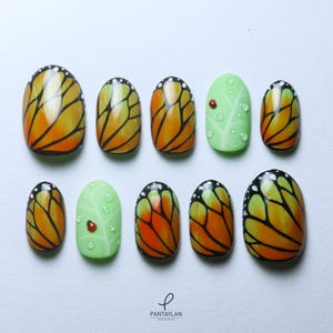 Könnte beinhalten: Satz mit zehn ovalen falschen Nägeln mit einem Schmetterlingsflügel-Design. Die Nägel sind in Orange, Schwarz und Weiß lackiert. Zwei der Nägel zeigen ein grünes Blatt mit einem roten Marienkäfer.