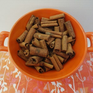 May include: An orange ceramic bowl filled with numerous cinnamon sticks. The bowl has two handles and sits on a patterned orange and white surface. The cinnamon sticks are brown and cylindrical.