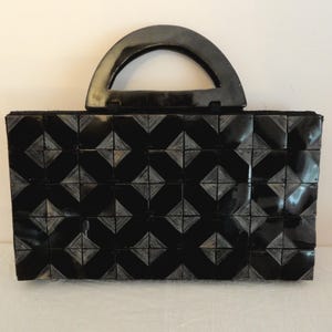 May include: A black handbag with a geometric pattern and a curved handle. The bag's body is made of small, square tiles arranged in a diamond pattern. The handle is a matching black color.