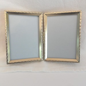 May include: A double-hinged picture frame with a metallic gold finish and a textured, repeating geometric pattern along the outer edges. The frame consists of two rectangular sections connected by a central metal hinge, holding blank white inserts.