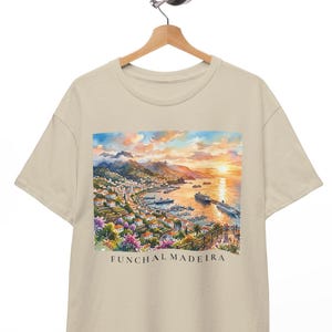 May include: A beige short-sleeve t-shirt featuring a colorful watercolor-style graphic print of the Funchal, Madeira coastline at sunset, including mountains, a harbor with cruise ships, and buildings. The text FUNCHAL MADEIRA is printed below.