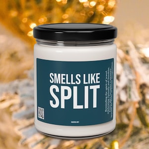 May include: A white jar candle with a black lid. The label features the text "SMELLS LIKE SPLIT" in white on a teal background. Additional text is present, along with a QR code. The candle is set against a blurred background with gold and red accents.