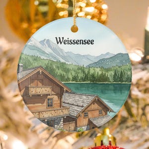 Könnte beinhalten: Runder Weihnachtsschmuck mit einem Aquarellgemälde eines Dorfes am See mit dem Text "Weissensee". Der Schmuck zeigt Holzhäuser, Berge und einen See, mit einem goldenen Faden zum Aufhängen.
