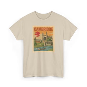 May include: Beige t-shirt featuring a vintage-style illustration of Cambridge, England. The design includes a bridge, buildings, a river, and a setting sun, with the word "CAMBRIDGE" at the top. The shirt is a neutral color.