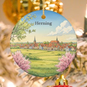 Puede incluir: Adorno navideño redondo de cerámica con una ilustración en acuarela de un paisaje urbano bajo un cielo azul. La palabra "Herning" está impresa en la parte superior. El adorno tiene una cuerda dorada para colgar y está decorado con una escena de la ciudad.