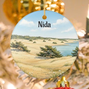 May include: Round ceramic ornament featuring a landscape scene with the word "Nida" in black text. The image depicts a sandy beach, a body of water, and trees under a blue sky with clouds. The ornament has a gold string for hanging.