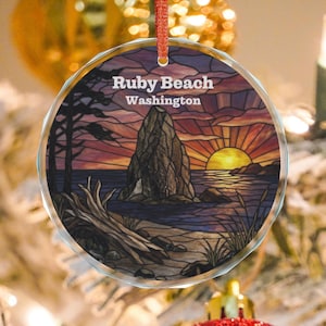 May include: A round, clear glass ornament featuring a stained-glass style print of Ruby Beach, Washington. The image depicts a sunset over the ocean, a large sea stack, driftwood on the sand, and pine trees. It hangs from a red ribbon.