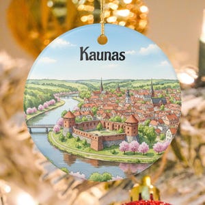 May include: Round ceramic ornament featuring a watercolor illustration of Kaunas, Lithuania. The artwork depicts a cityscape with a river, castle, and buildings under a blue sky. The ornament has a gold string for hanging.