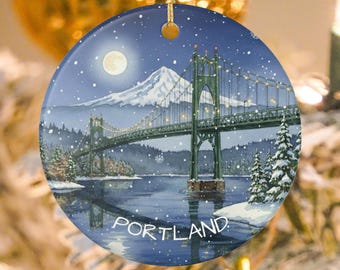 Könnte beinhalten: Runder Weihnachtsbaumschmuck aus Keramik mit einer Winterlandschaft einer grünen Brücke über Wasser und einem schneebedeckten Berg im Hintergrund. Das Wort "PORTLAND" ist unten aufgedruckt. Der Schmuck hängt an einem goldenen Faden.