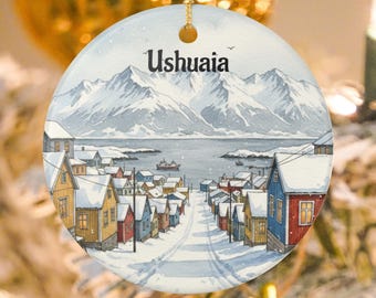 Könnte beinhalten: Runder Weihnachtsschmuck mit einer Aquarellillustration von Ushuaia, Argentinien. Das Design zeigt ein verschneites Dorf am Fuße schneebedeckter Berge, mit einem Gewässer und einem Schiff im Hintergrund. Das Wort "Ushuaia" ist oben aufgedruckt.