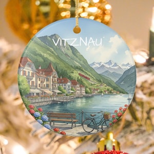 May include: Round ceramic ornament featuring a watercolor painting of Vitznau, Switzerland. The image shows a lakeside town with buildings, mountains, a lake, and a bicycle. The word "VITZNAU" is printed at the top.