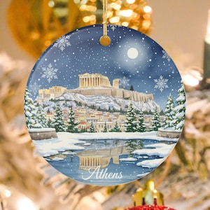 May include: Round Christmas ornament featuring a winter scene of Athens, Greece, with the Parthenon on a snowy hill. The ornament has a blue background with snowflakes and a full moon. The word "Athens" is written at the bottom.