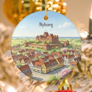 May include: Round ceramic Christmas ornament featuring a watercolor illustration of Nyborg, Denmark. The image depicts a castle, town, and surrounding landscape under a blue sky. The word "Nyborg" is written across the top.