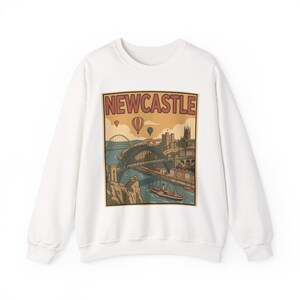 May include: White sweatshirt featuring a vintage-style illustration of Newcastle, England. The graphic includes a bridge, buildings, hot air balloons, and the word "NEWCASTLE" in a retro font. A cozy, casual top.