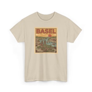 May include: A beige t-shirt featuring a rectangular graphic with the word "BASEL" at the top. The graphic depicts a detailed illustration of a city with a river, buildings, and a setting sun. The shirt is made of a soft material.