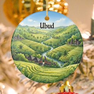 Puede incluir: Adorno redondo de cerámica con una pintura de acuarela de Ubud, Bali. La obra representa exuberantes terrazas de arroz verdes, un río y edificios tradicionales bajo un cielo azul. La palabra "Ubud" está escrita en la parte superior.