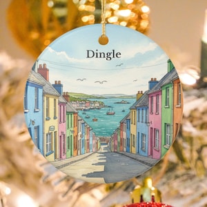 May include: Round ceramic ornament featuring a watercolor painting of Dingle, Ireland. The artwork depicts a street lined with colorful buildings leading to a bay with boats. The word "Dingle" is printed at the top.