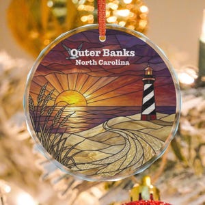Puede incluir: Un adorno redondo de cristal transparente con una ilustración estilo vidriera de un faro en una playa de arena al atardecer. El texto Outer Banks North Carolina está impreso en blanco. Una cinta roja está sujeta en la parte superior.